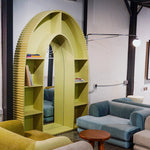 تحميل الصورة إلى عارض المعرض، A contemporary, arch-shaped bookshelf in a vibrant yellow color with integrated mirror, situated in a modern living space. The bookshelf features grooved edges and contains books and decorative items. The area includes comfortable blue and beige sofas and a small wooden table, creating a cozy and stylish ambiance. Buy Bookshelf Dubai